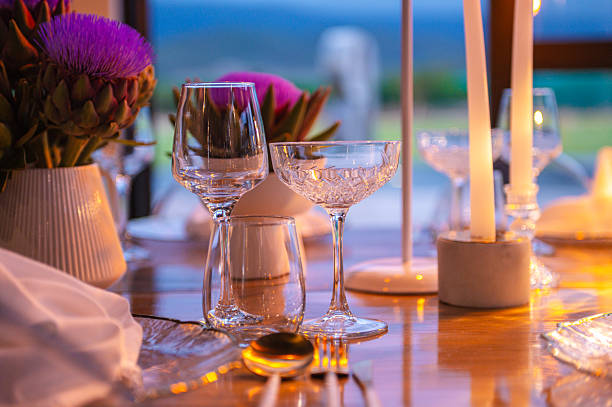 Elegant candlelit dinner setting at dusk featuring wine glasses, a crystal coupe, soft candlelight and purple floral centerpieces on a polished wooden table. Warm, intimate atmosphere ideal for romantic dinners, special celebrations or upscale restaurant scenes with refined tableware and gentle bokeh background.